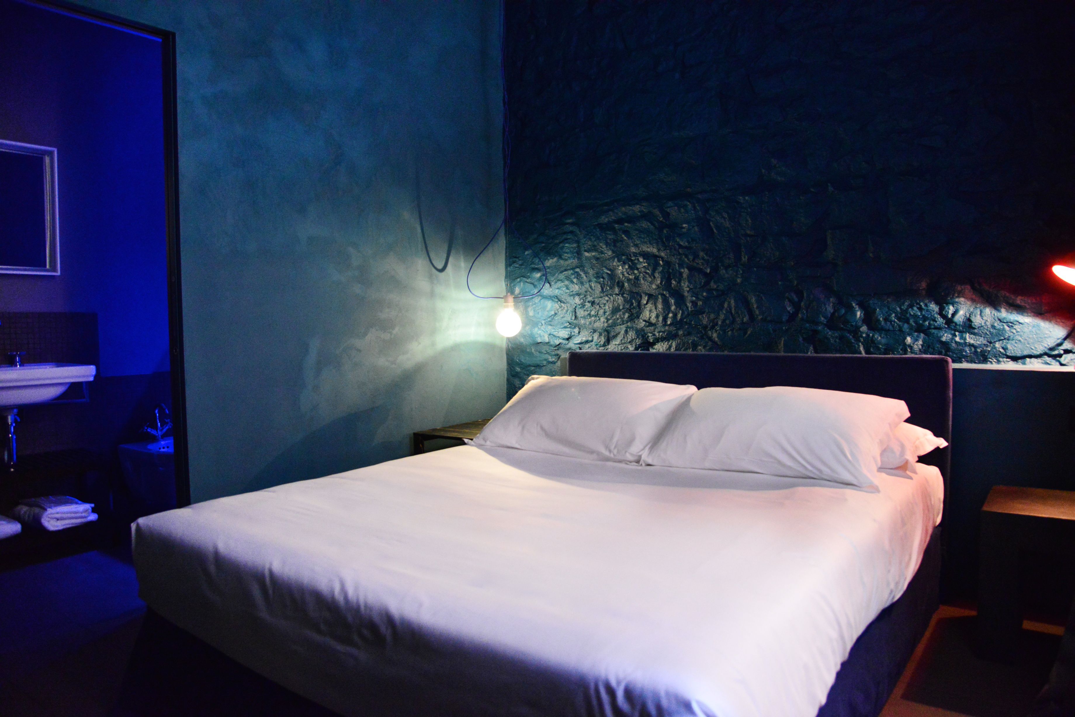 A neatly made bed with white linens in a dimly lit room with a textured wall and a small nightstand with a lamp.