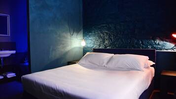 A neatly made bed with white linens in a dimly lit room with a textured wall and a small nightstand with a lamp.