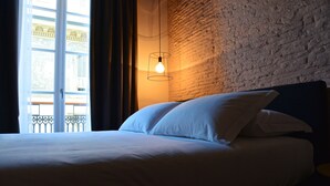 Een bed met witte lakens in een kamer met een bakstenen muur en een raam met gordijnen.