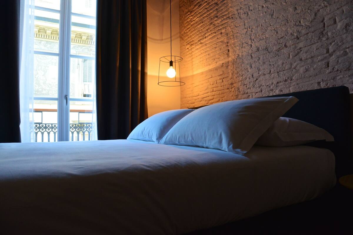 Una cama con sábanas blancas en una habitación con pared de ladrillo y ventana con cortinas.