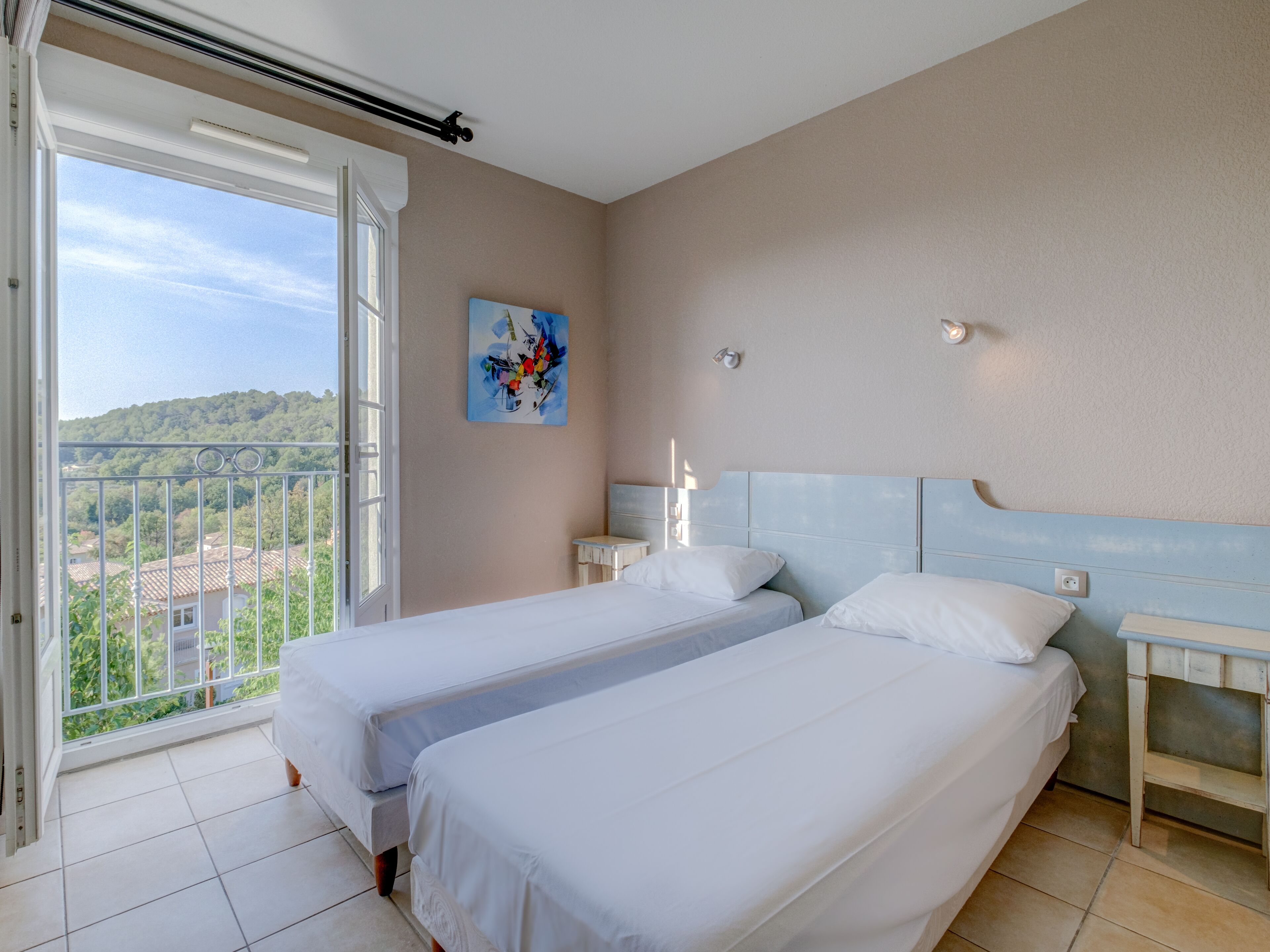 A hotel room with two beds, a balcony with a view of the city, and a painting on the wall.
