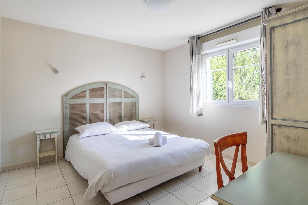Un dormitorio con una cama, una silla de madera, una mesita, una ventana y una puerta.