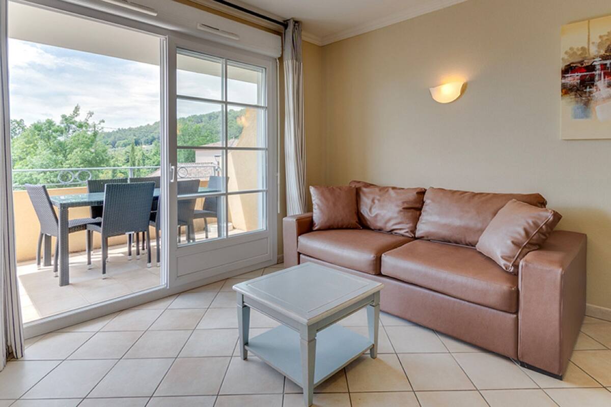 Una sala de estar con un sofá de cuero marrón, una mesa de centro blanca y un ventanal corredizo que da a un patio exterior con mesa y sillas.