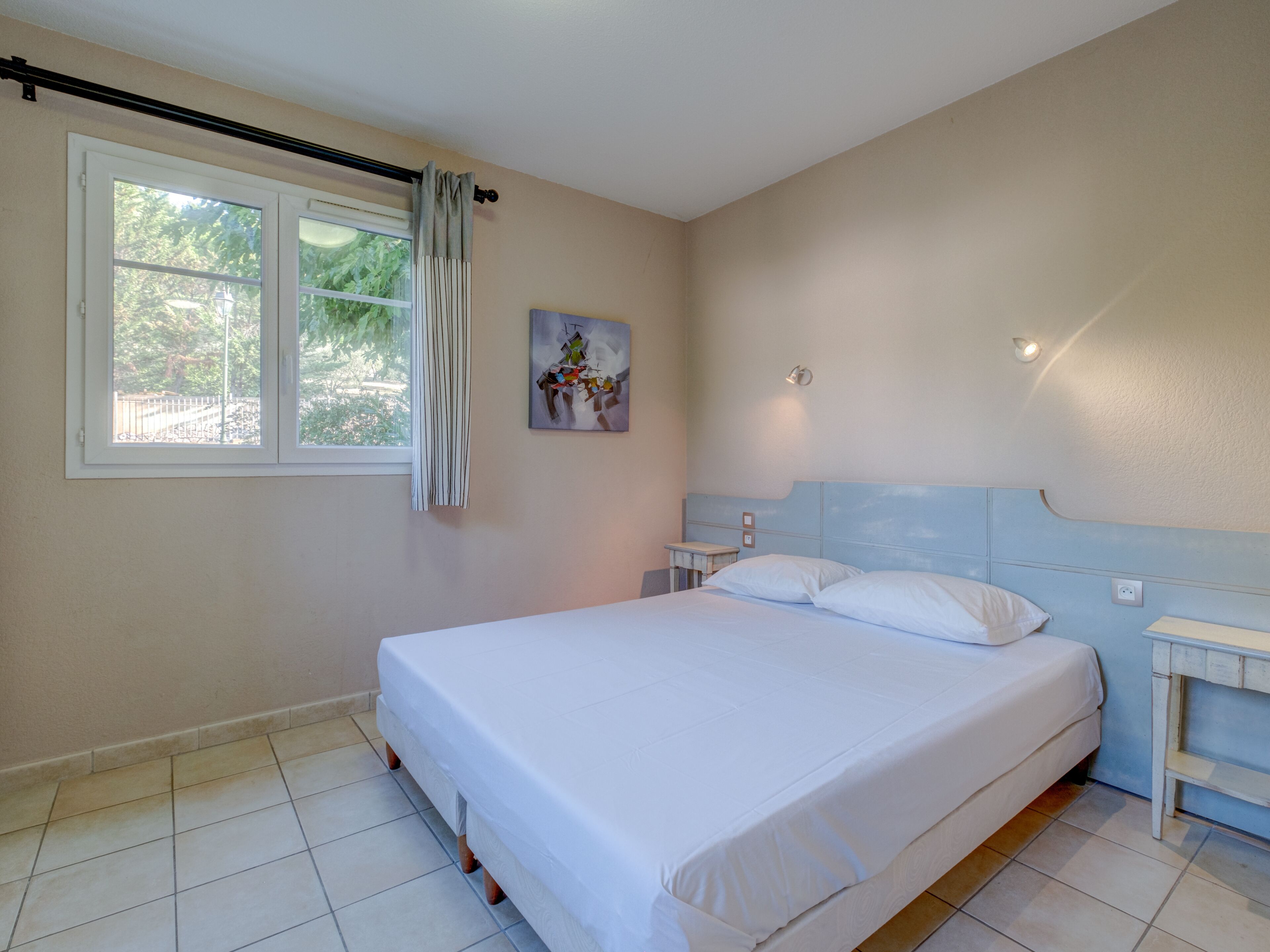 A bedroom with a bed, a nightstand, a window with curtains, and a wall-mounted light.