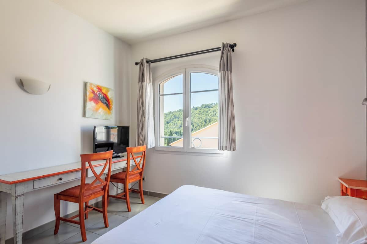 Un dormitorio con cama, escritorio con silla, televisor y ventana con cortinas.
