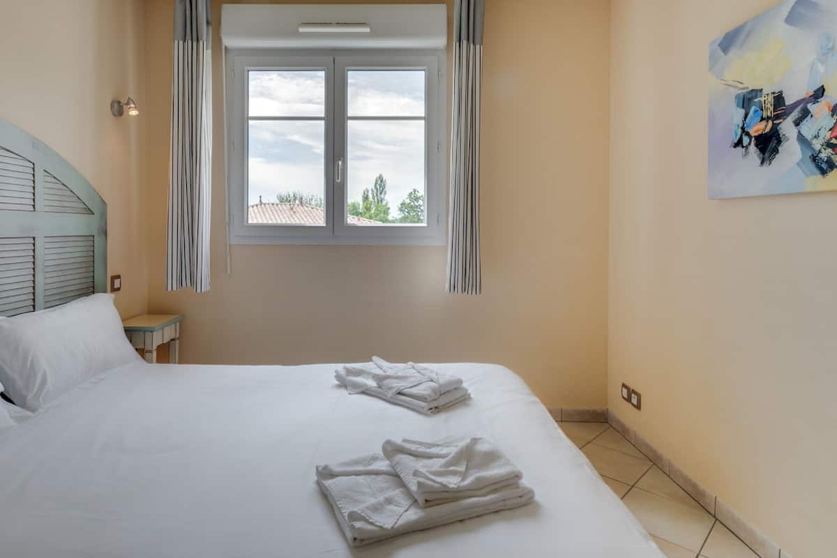 Un dormitorio con cama, mesita de noche, ventana con cortinas y un cuadro en la pared.