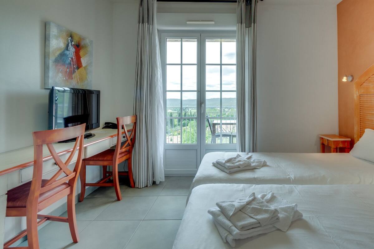 Habitación de hotel con dos camas, un escritorio con silla, televisor y balcón con vistas.