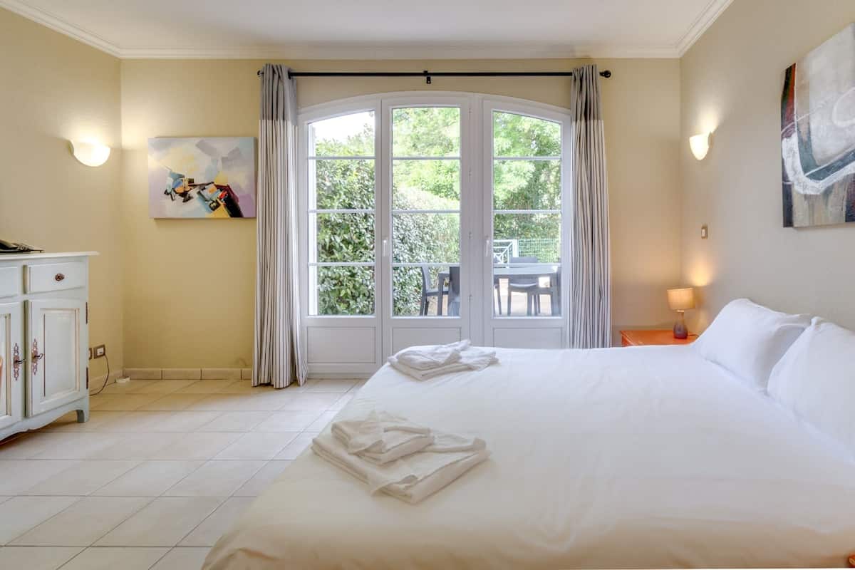 Habitación de hotel con una cama grande, zona de comedor con sillas y una mesa, y vistas a zonas verdes.
