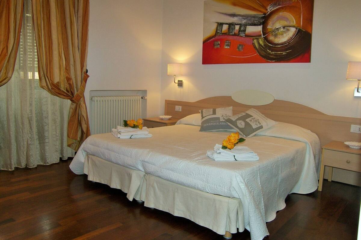 A hotel room with a large bed, a radiator, a painting on the wall, and a window with curtains.