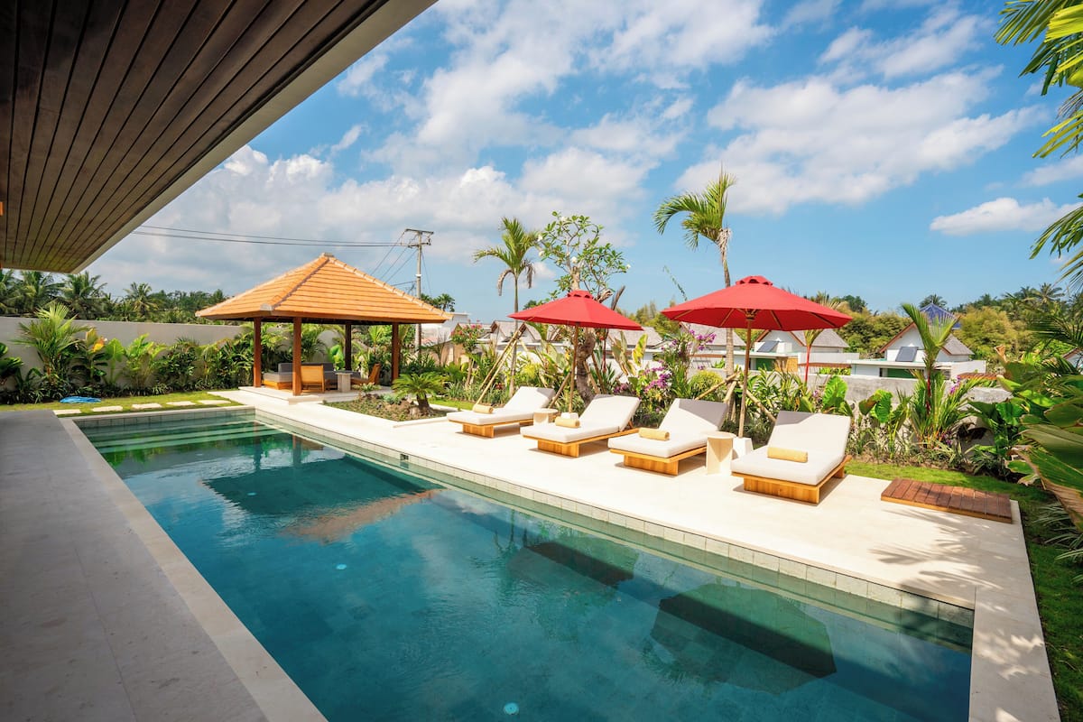 Piscina com espreguiçadeiras e guarda-sóis vermelhos, cercada por plantas tropicais e um gazebo de madeira.