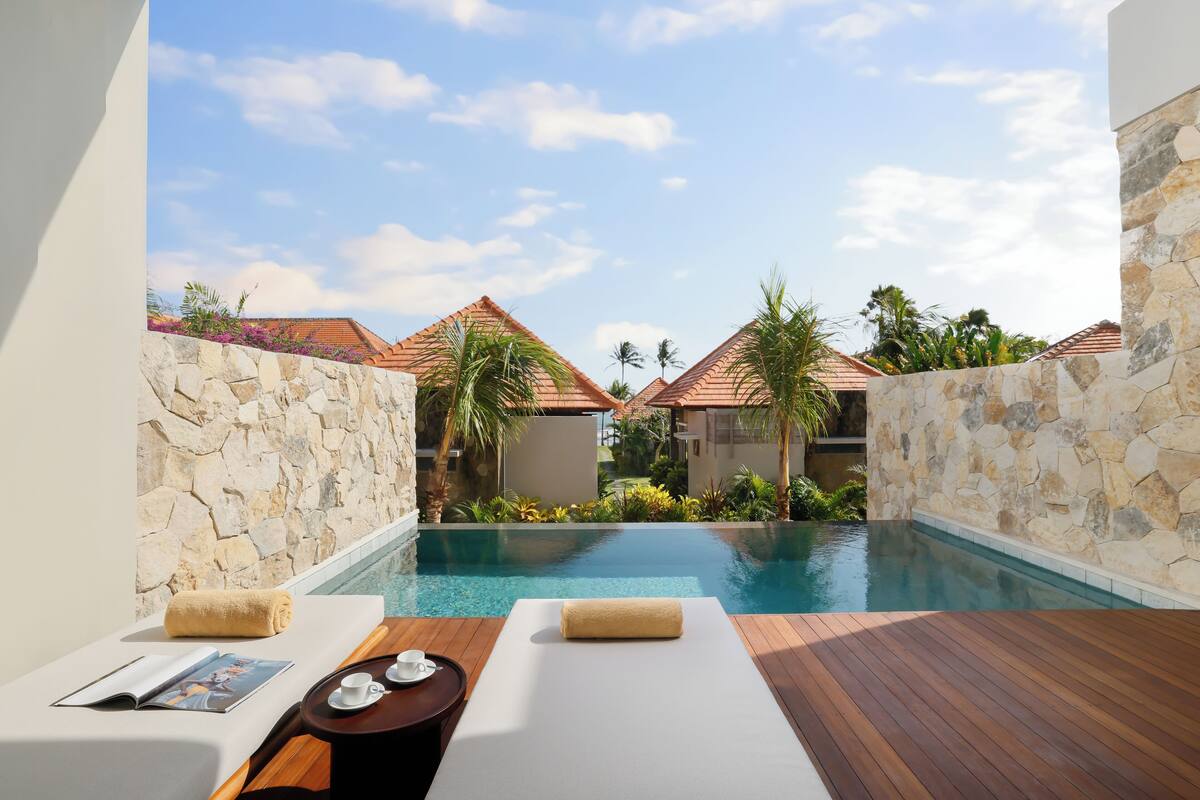 Uma área de relaxamento à beira da piscina com um deck de madeira, uma parede de pedra e vista para um resort com telhados em azulejo.