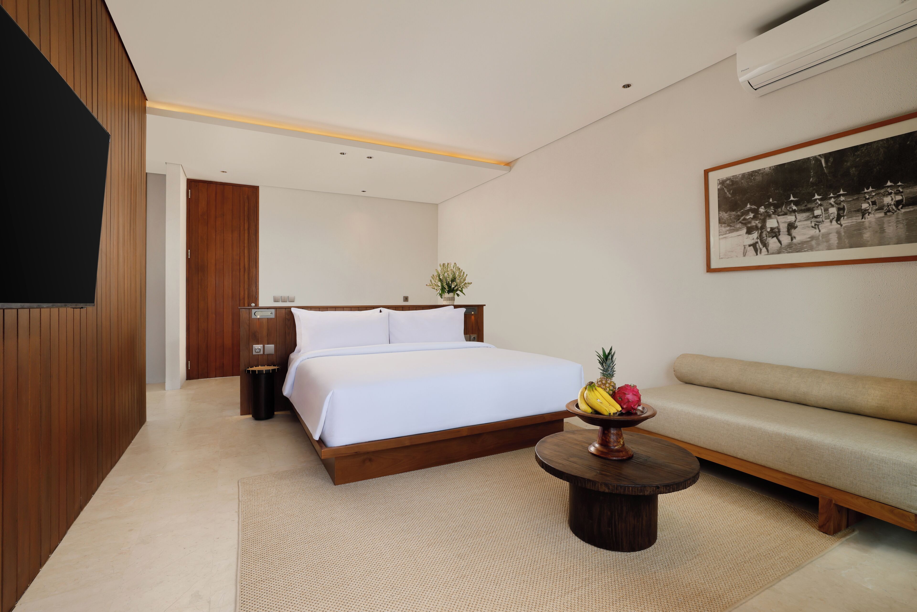 Quarto de hotel moderno com uma cama, um sofá, uma mesa de centro com frutas, uma TV fixada na parede e um quadro emoldurado.