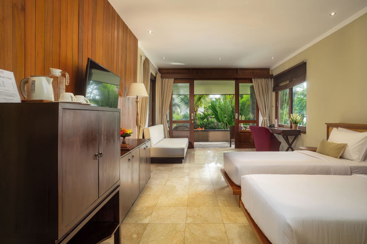 Quarto de hotel com armário de madeira, televisão, escrivaninha com cadeira, duas camas e vista para o verde lá fora.