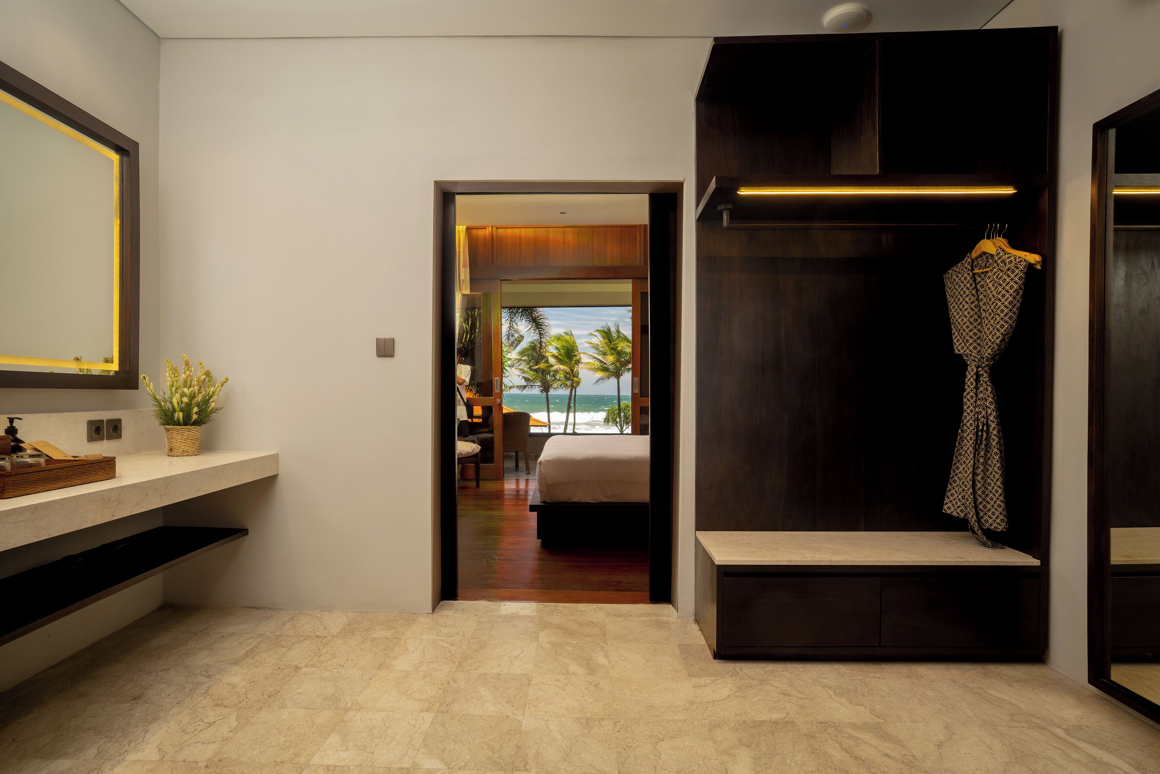 A modern hotel room with a marble floor, a large mirror, a bedside table, a wardrobe, and a view of the ocean.