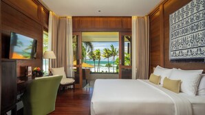 Habitación de hotel con una cama grande, un escritorio, una silla y un televisor. Se ve una vista de palmeras y el océano a través de las ventanas.