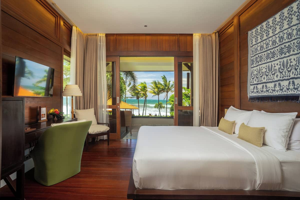 Quarto de hotel com uma cama grande, uma mesa, uma cadeira e uma televisão. As janelas oferecem vista para palmeiras e o oceano.