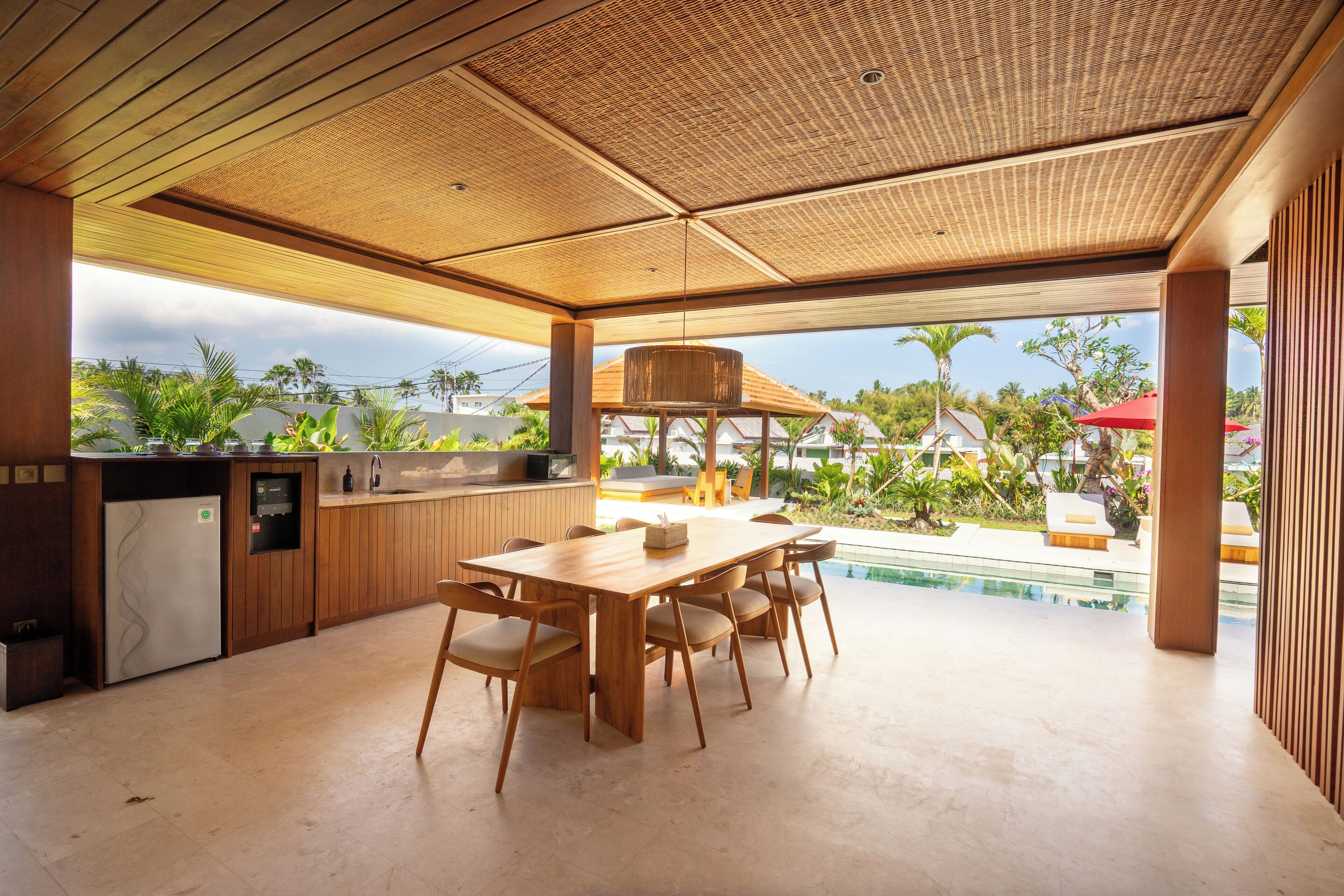 A modern kitchen with a wooden table and chairs, a built-in refrigerator, and a view of a swimming pool and tropical plants.