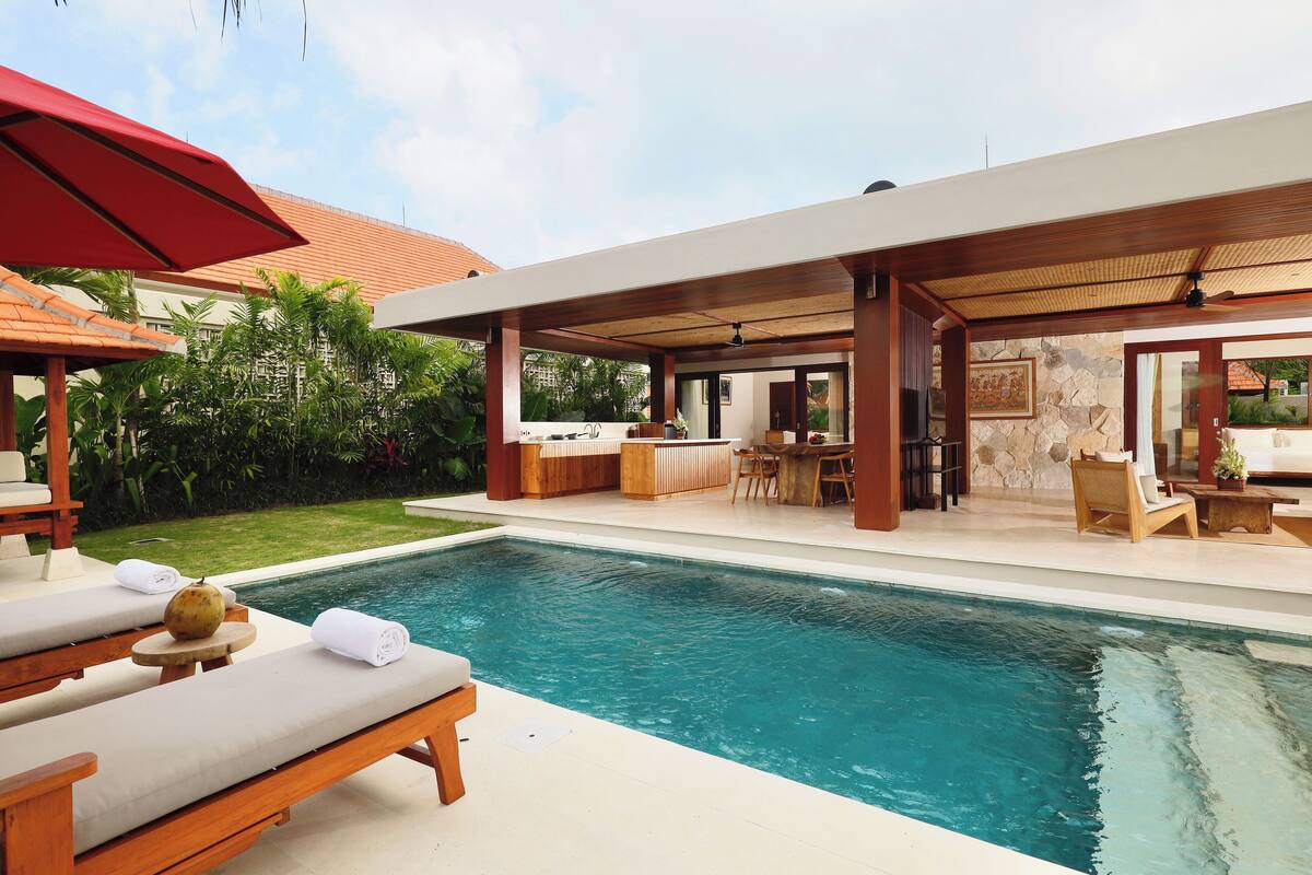 Uma área junto à piscina com espreguiçadeiras, bar e espaço coberto para sentar.