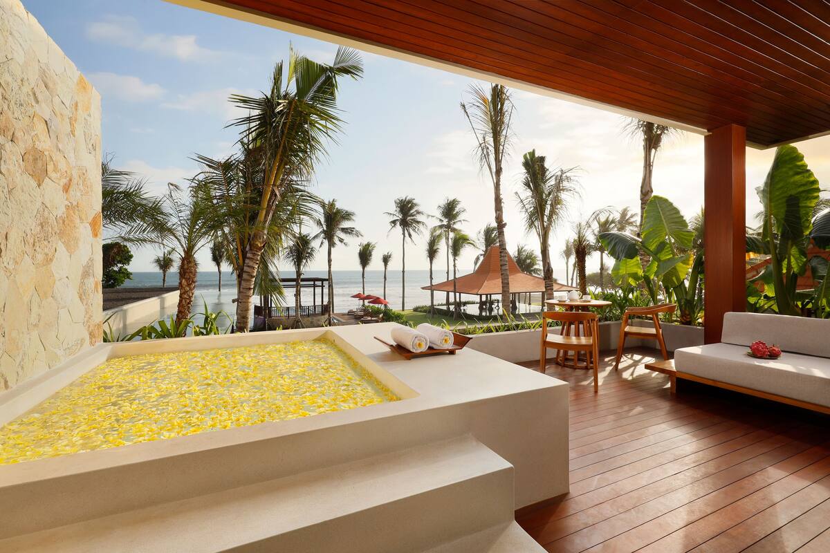 Uma área de spa com uma banheira grande cheia de flores amarelas, assentos de deck de madeira e vista para a praia e palmeiras.