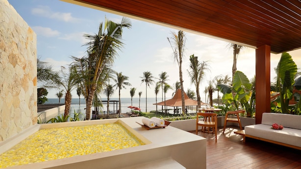 Un área de spa con una bañera grande llena de flores amarillas, asientos de madera en terraza y vistas a la playa y palmeras.