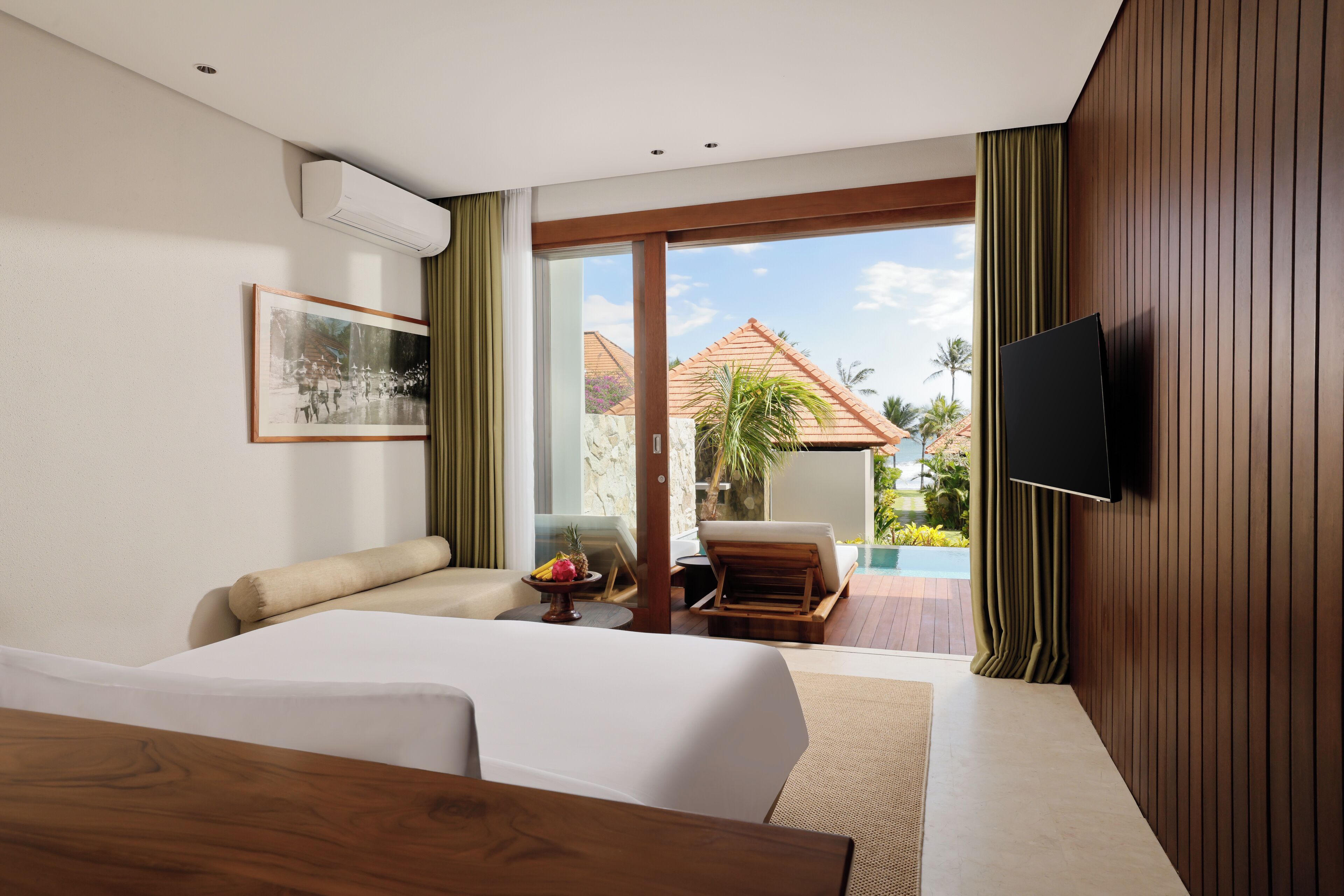 Une chambre d’hôtel moderne avec un grand lit, un téléviseur à écran plat, une petite table avec des fruits, et une vue sur une piscine et des palmiers.