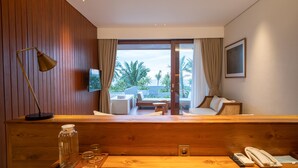 Una habitación de hotel moderna con un mostrador de recepción de madera, televisión, un ventanal con vistas a palmeras y una playa, y un rincón de descanso acogedor con sofá y una mesita.