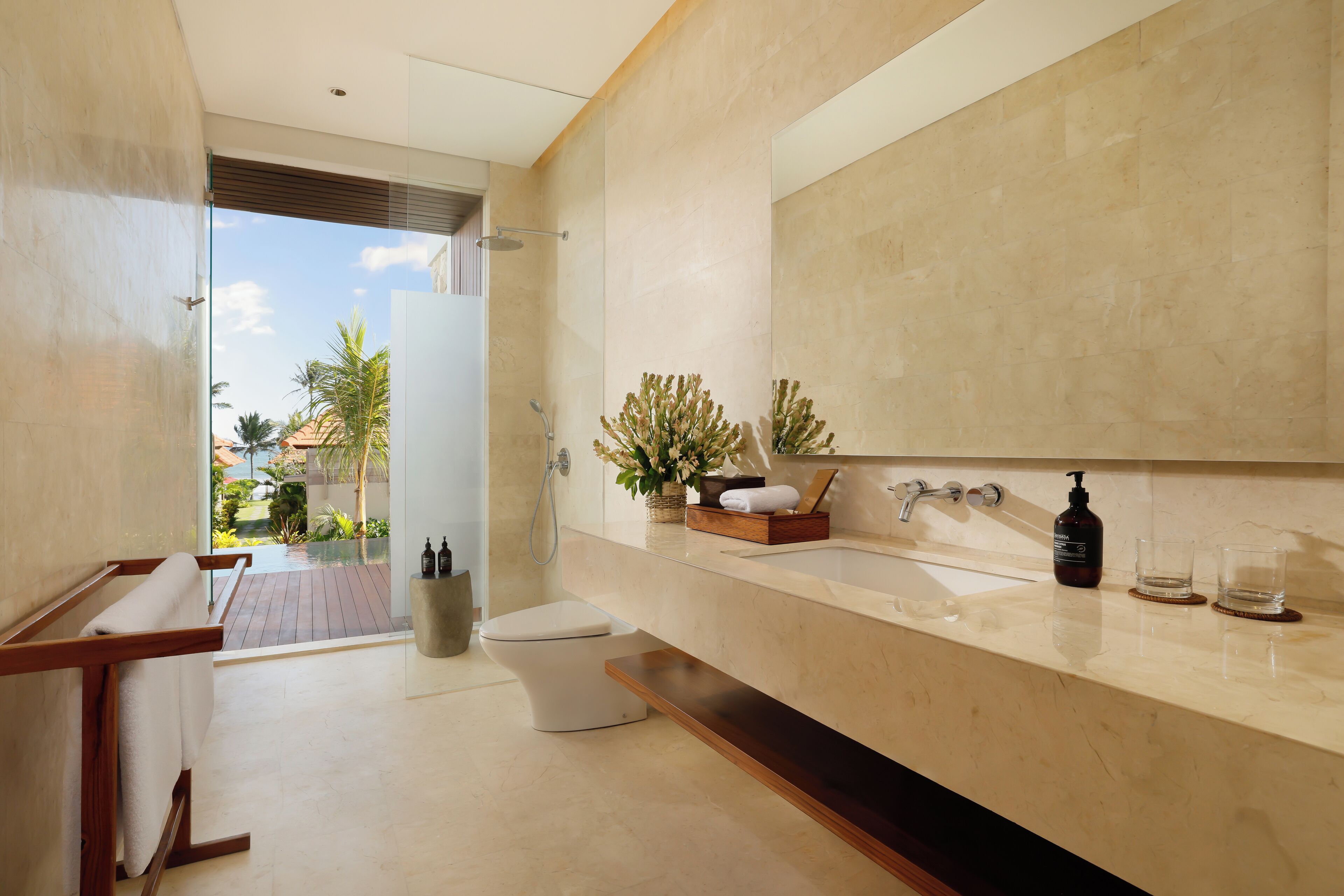 A modern bathroom with a large mirror, a bathtub, and a toilet. There is a window providing a view of palm trees and a garden.