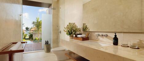 A modern bathroom with a large mirror, a bathtub, and a toilet. There is a window providing a view of palm trees and a garden.