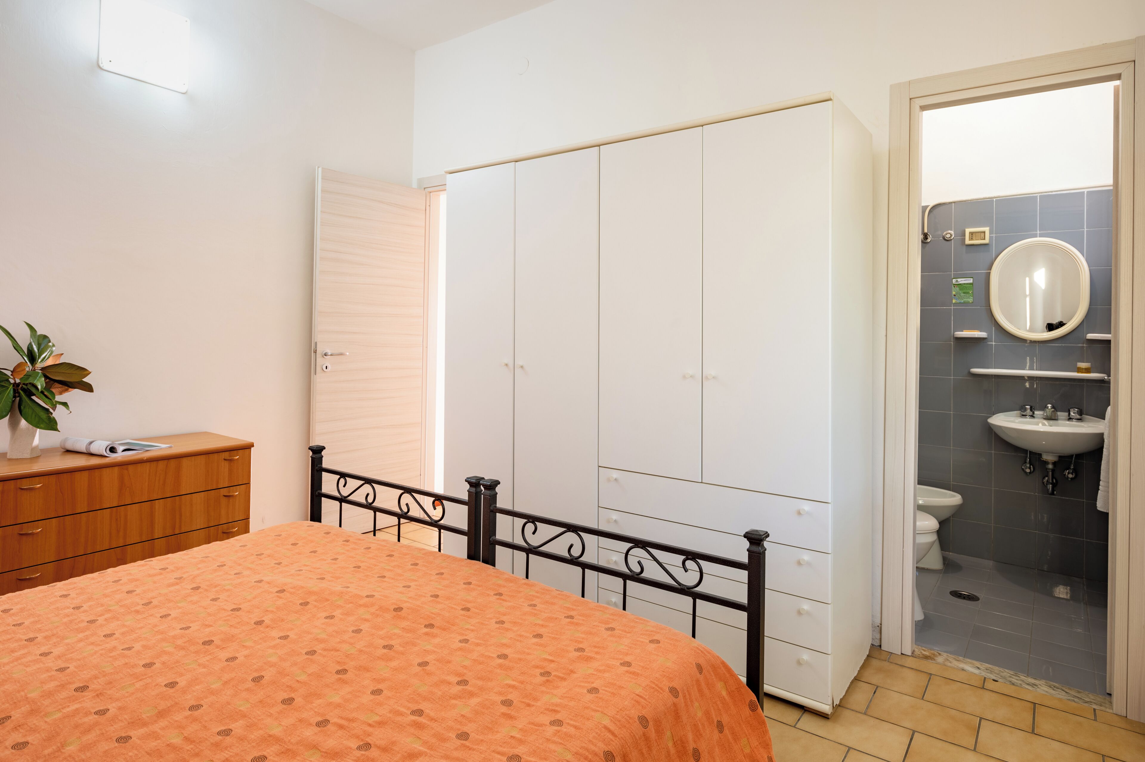 A bedroom with a bed, a wardrobe, a bedside table, and a bathroom with a sink and toilet.