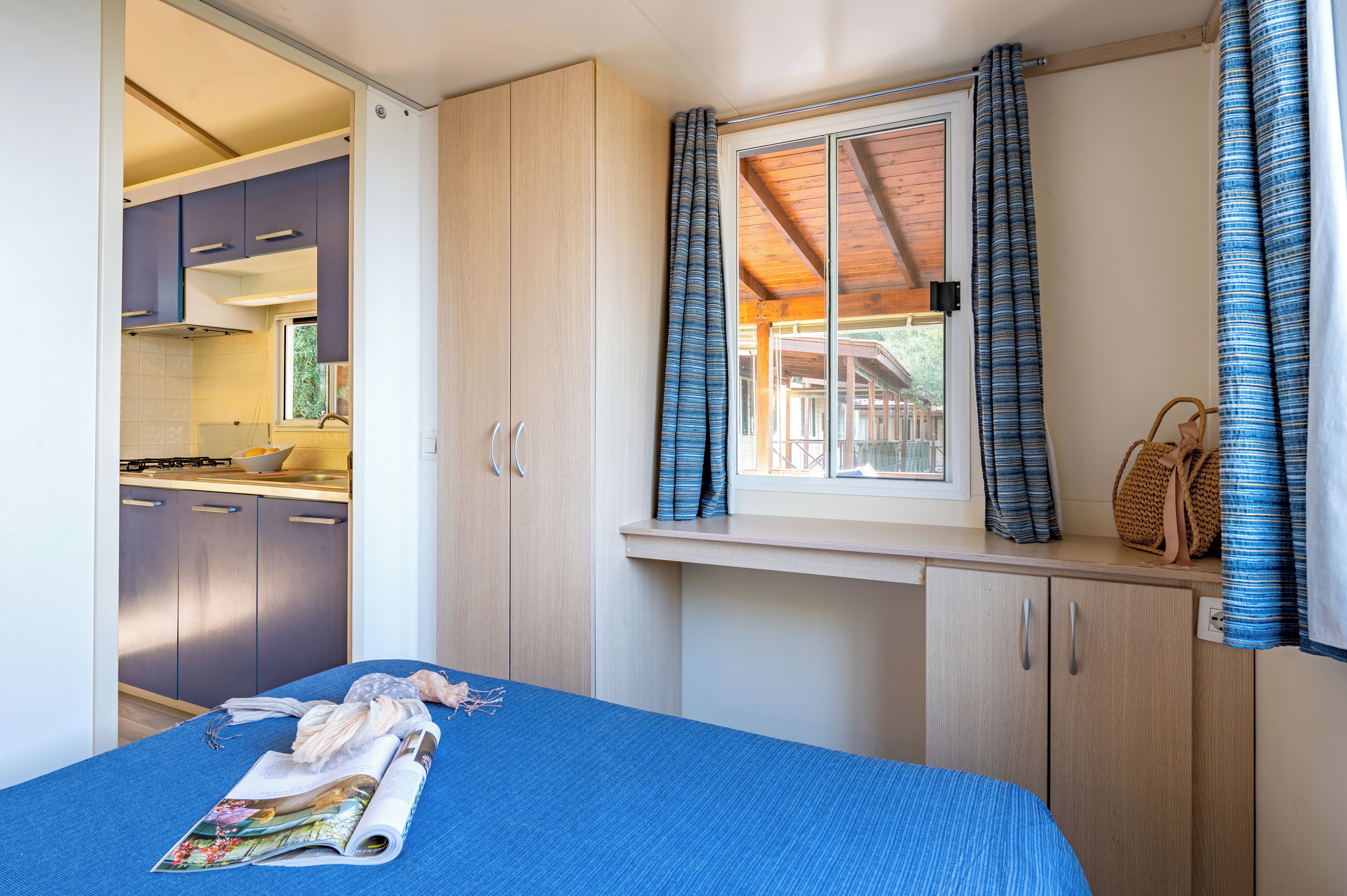 Un dormitorio compacto con una cama individual, un ventanal con cortinas y una kitchenette visible a través de una puerta abierta.