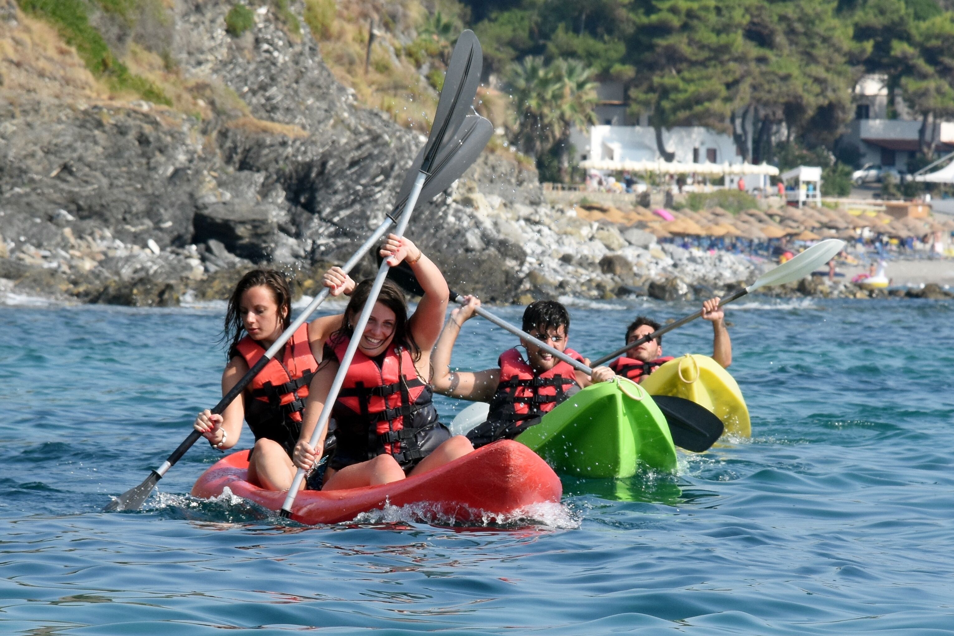 Un groupe de personnes se trouve dans un kayak rouge et vert sur une étendue d’eau, avec une rive rocheuse et des bâtiments en arrière-plan.