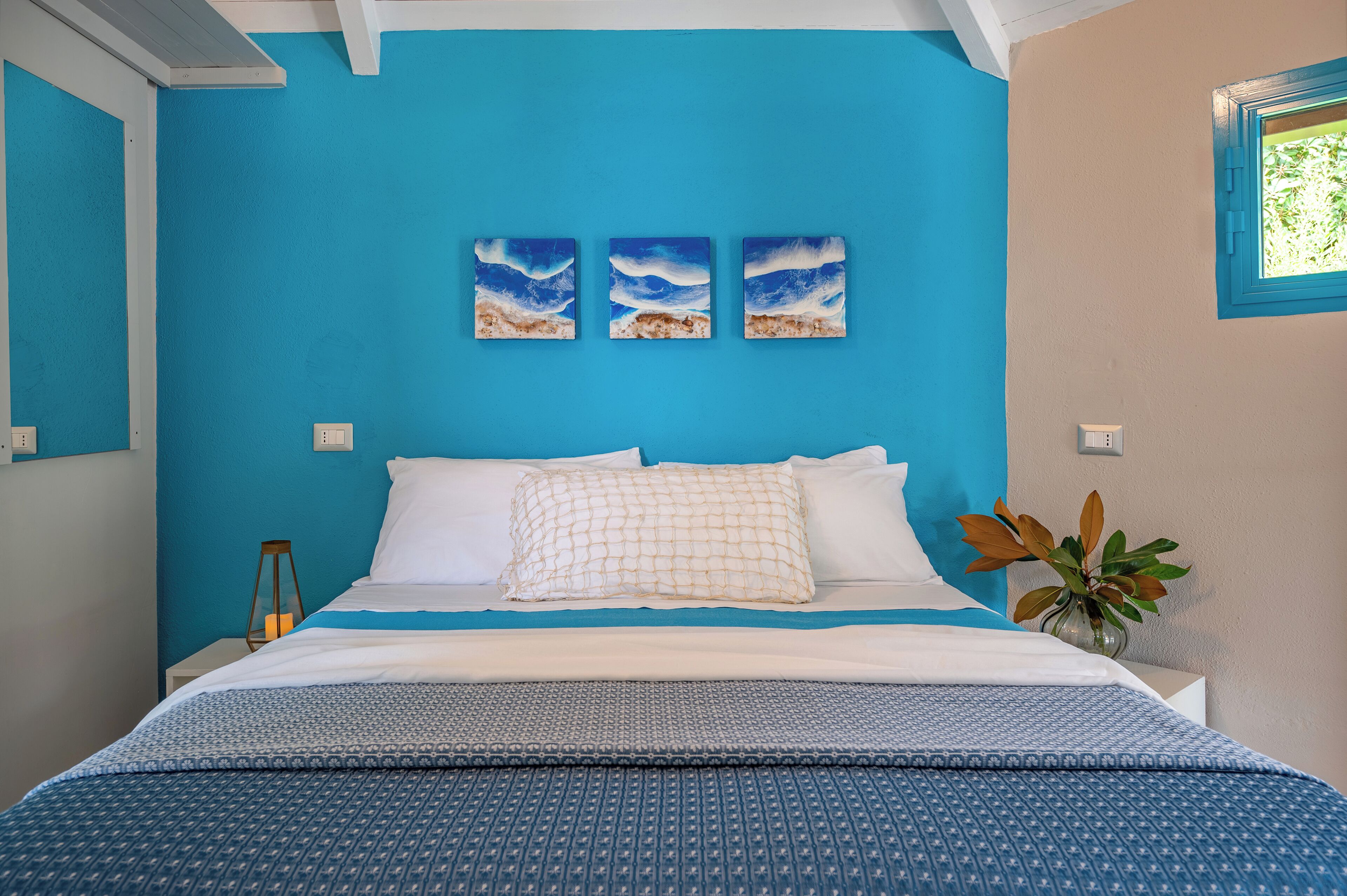 Ein Schlafzimmer mit blauer Wand, einem Bett mit weißer und gemusterter Bettwäsche, drei Wandbildern, einem Nachttisch mit Lampe und einer Topfpflanze.