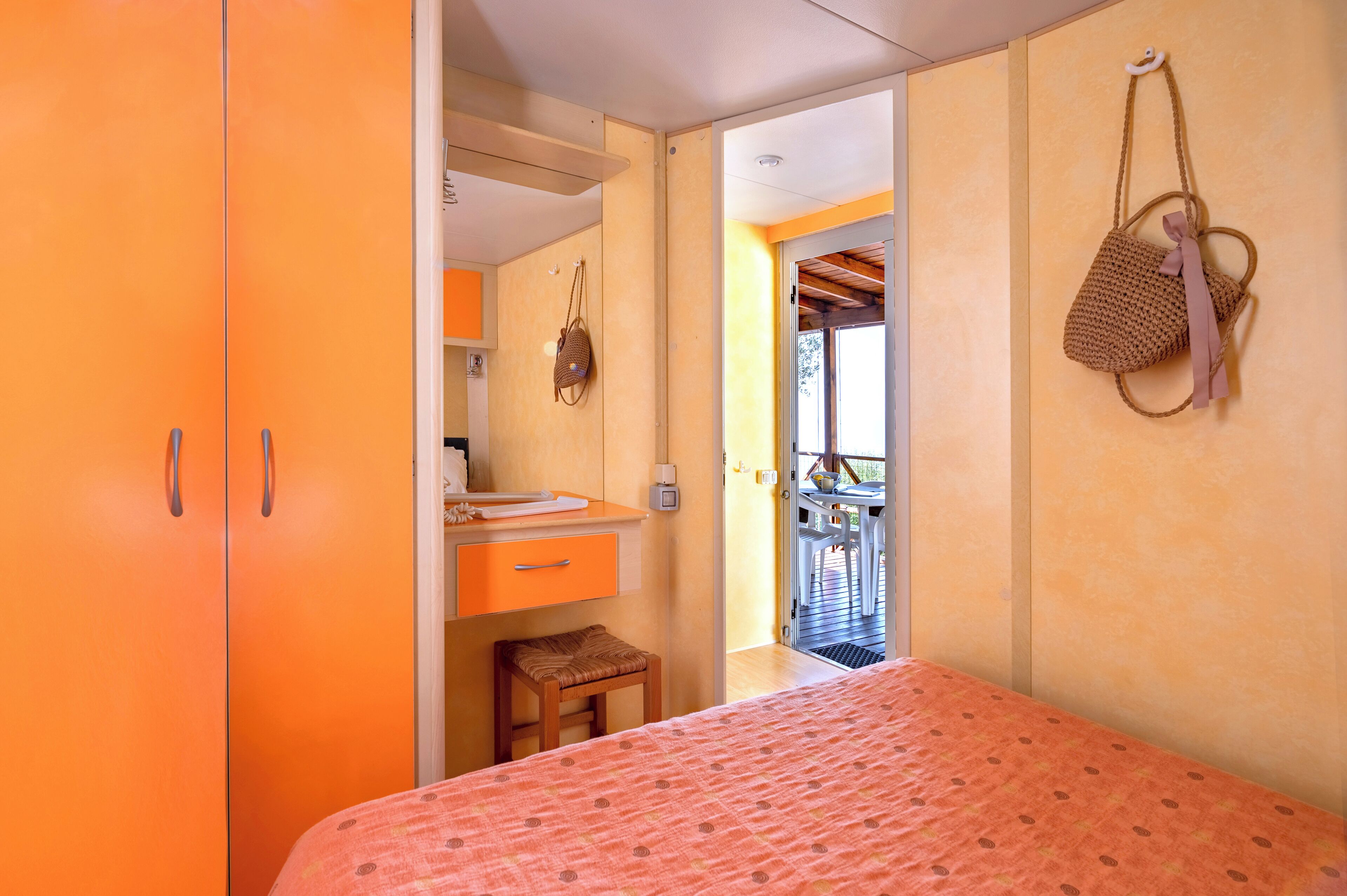 A bedroom with an orange wardrobe, a small wooden stool, a bed with a patterned bedspread, and a view of an outdoor balcony.