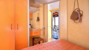 A bedroom with an orange wardrobe, a small wooden stool, a bed with a patterned bedspread, and a view of an outdoor balcony.