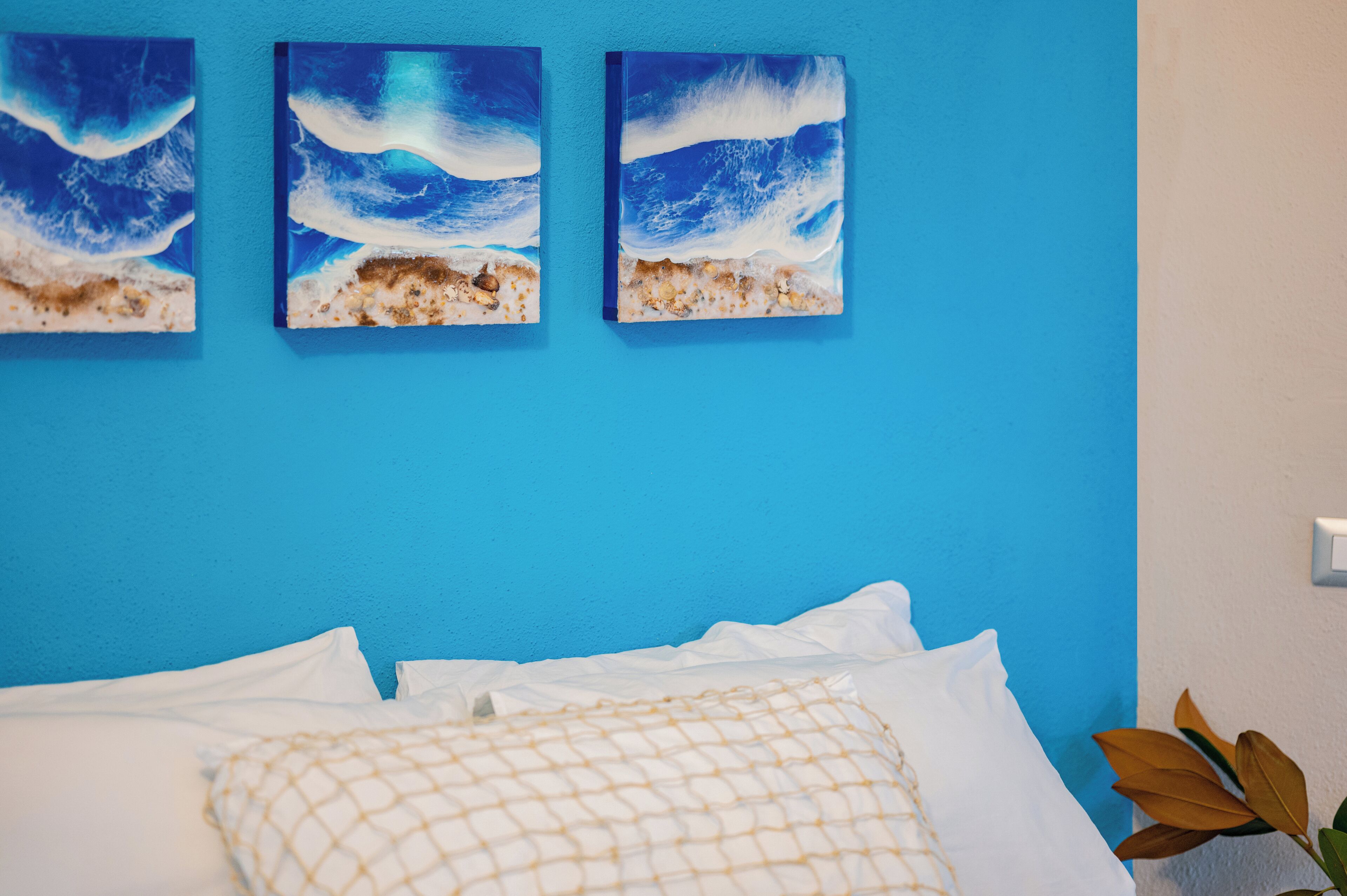 A bed with white linens and a beige netted pillow against a blue wall with three ocean wave paintings.