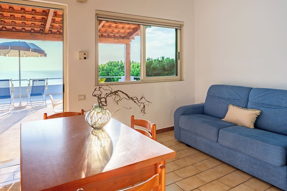 Ein Zimmer mit Esstisch, Stühlen, einem blauen Sofa, einem Fenster mit Blick auf Grünflächen und einem Deckenventilator.