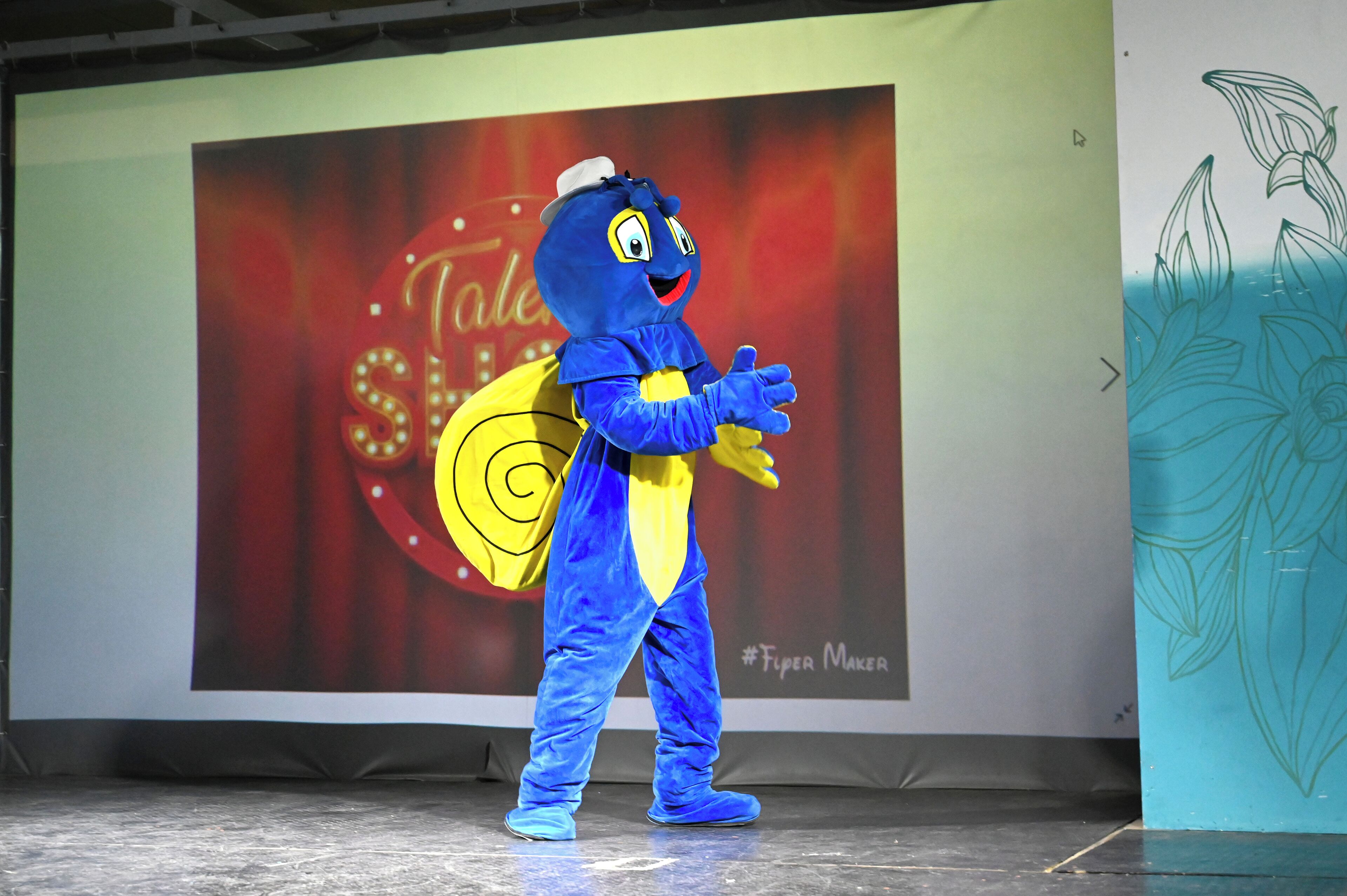 A mascot in a blue and yellow costume on a stage with a large screen in the background displaying the word "Tale" and a hashtag "#TiferMaker".