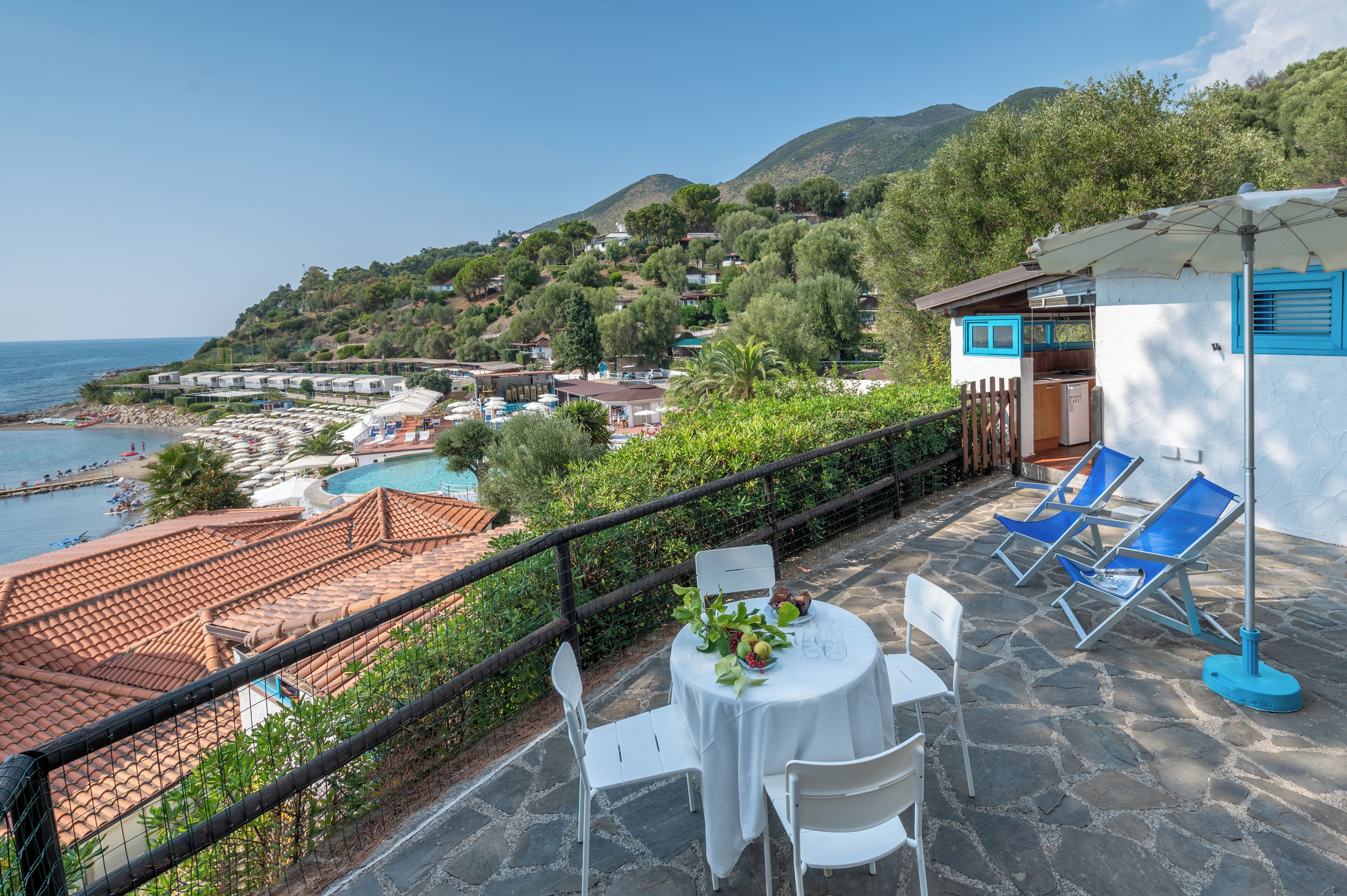 Una terrazza con un tavolo apparecchiato per mangiare, con vista su una zona costiera che include una piscina e una spiaggia.