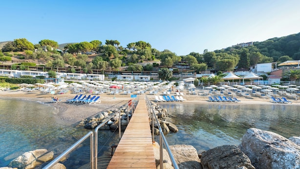 Une jetée en bois qui mène à une plage où s’alignent des rangées de chaises longues et de parasols.