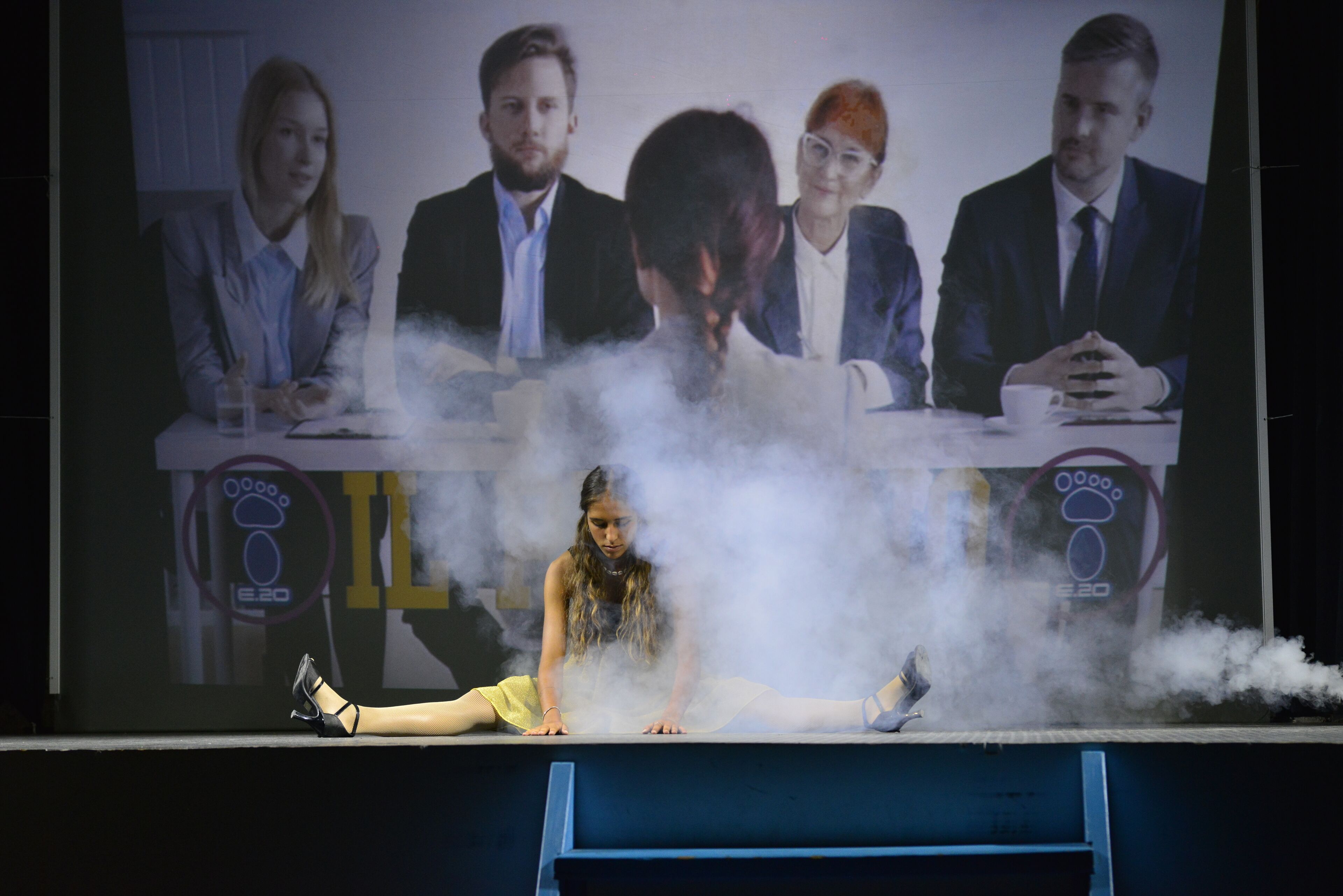 A stage with a person lying on the floor surrounded by smoke, with a group of people in the background.