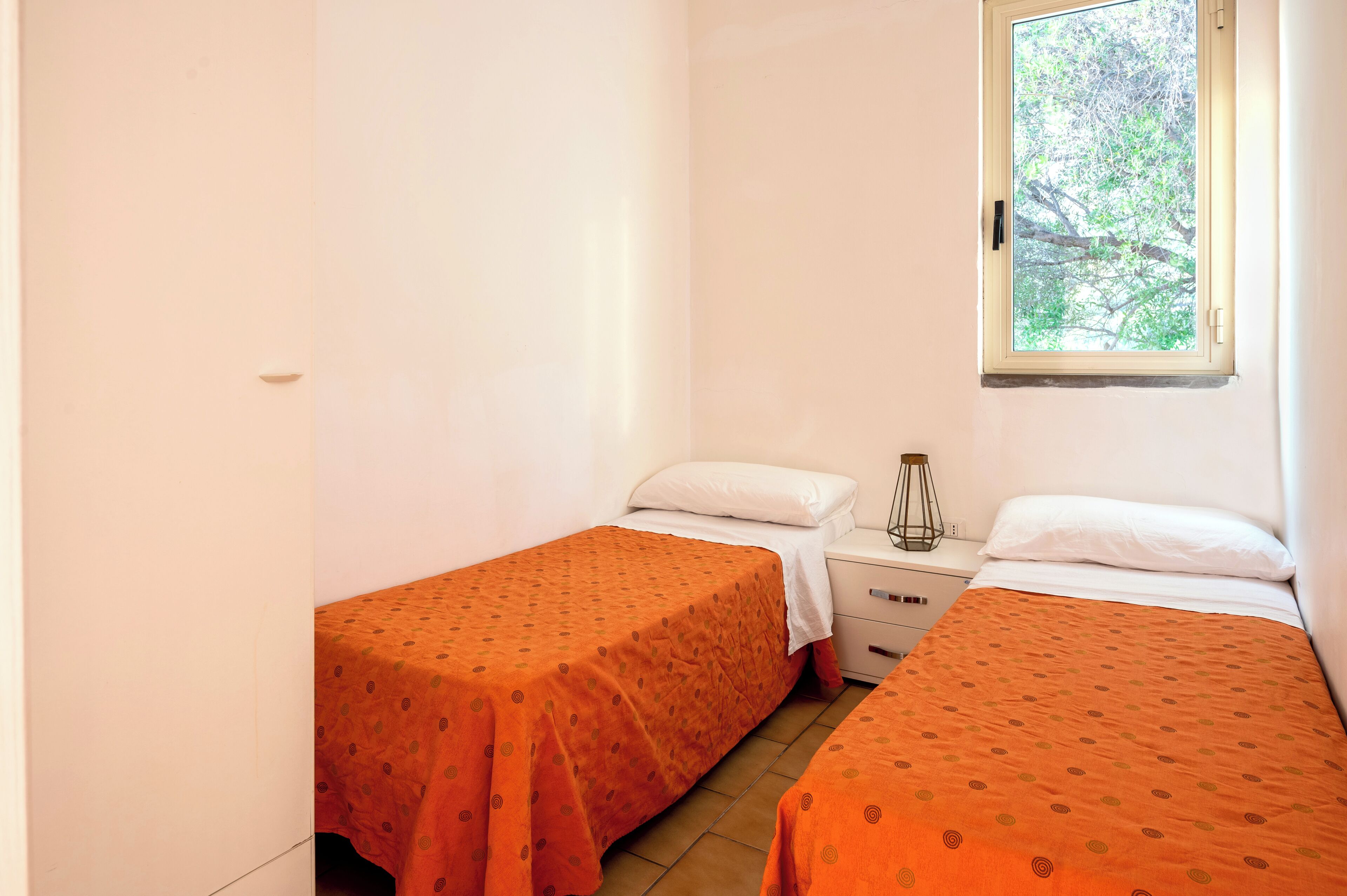 A small, clean room with two single beds, each with an orange bedspread, a bedside table with a lamp, and a window with a view of trees.