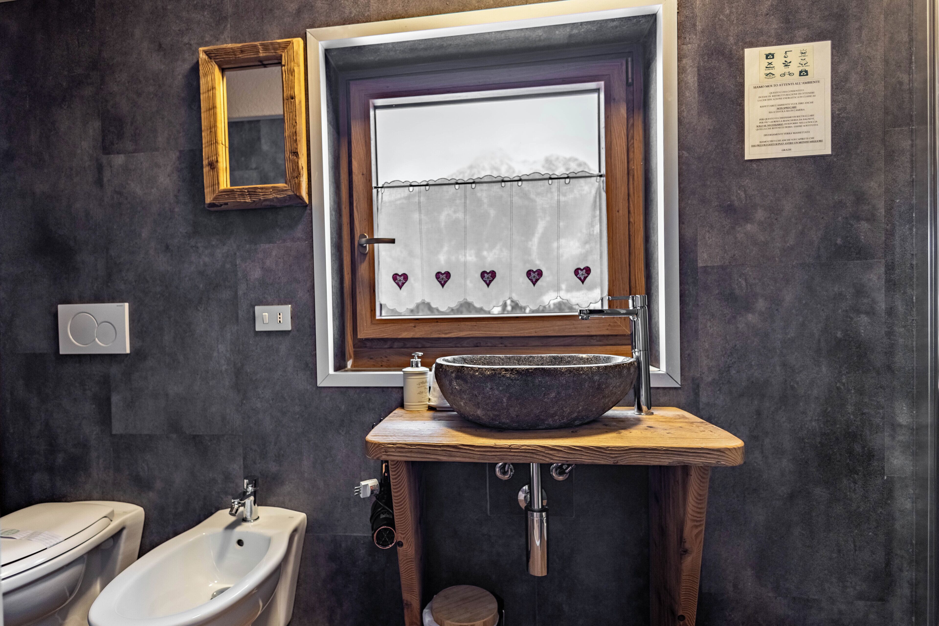 A bathroom with a large window, a wooden vanity, a white bidet, and a stone sink.