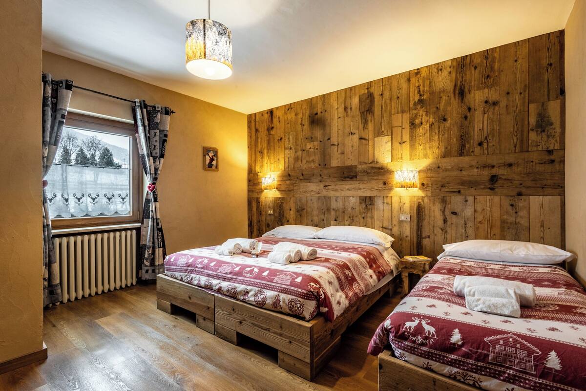 Une pièce chaleureuse aux murs en bois, avec deux lits dotés de draps rouges et blancs, une fenêtre avec des rideaux et un radiateur.