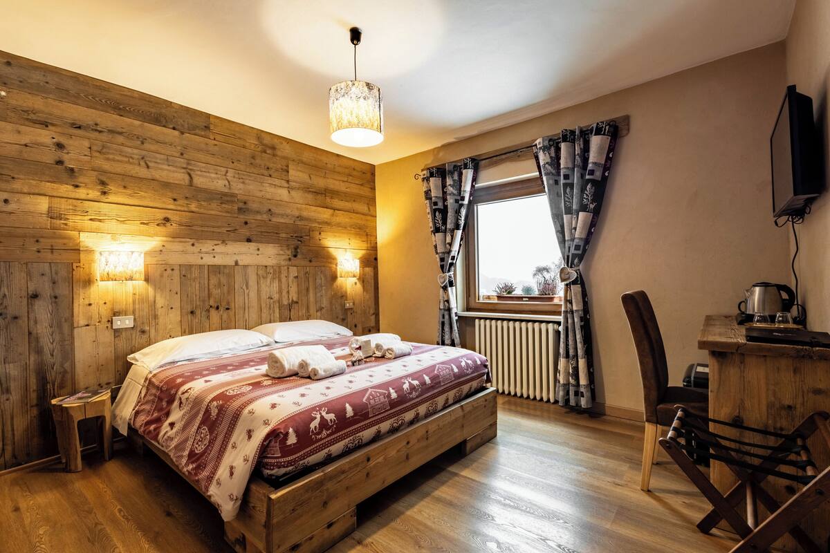 Une chambre à coucher aux murs lambrissés, comprenant un lit, des lampes de chevet, une fenêtre avec des rideaux, un radiateur et un bureau avec une bouilloire.