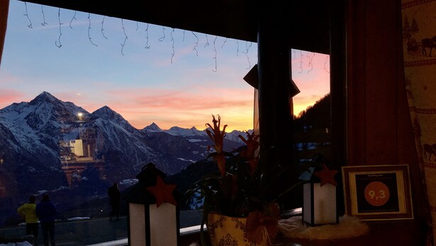 Una habitación con vistas a montañas nevadas al atardecer, una ventana con adornos colgantes, y una mesa con un farol y un diploma enmarcado.