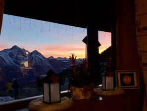 Ein Zimmer mit Blick auf verschneite Berge bei Sonnenuntergang, ein Fenster mit hängenden Dekorationen und ein Tisch mit Laterne und gerahmtem Zertifikat.