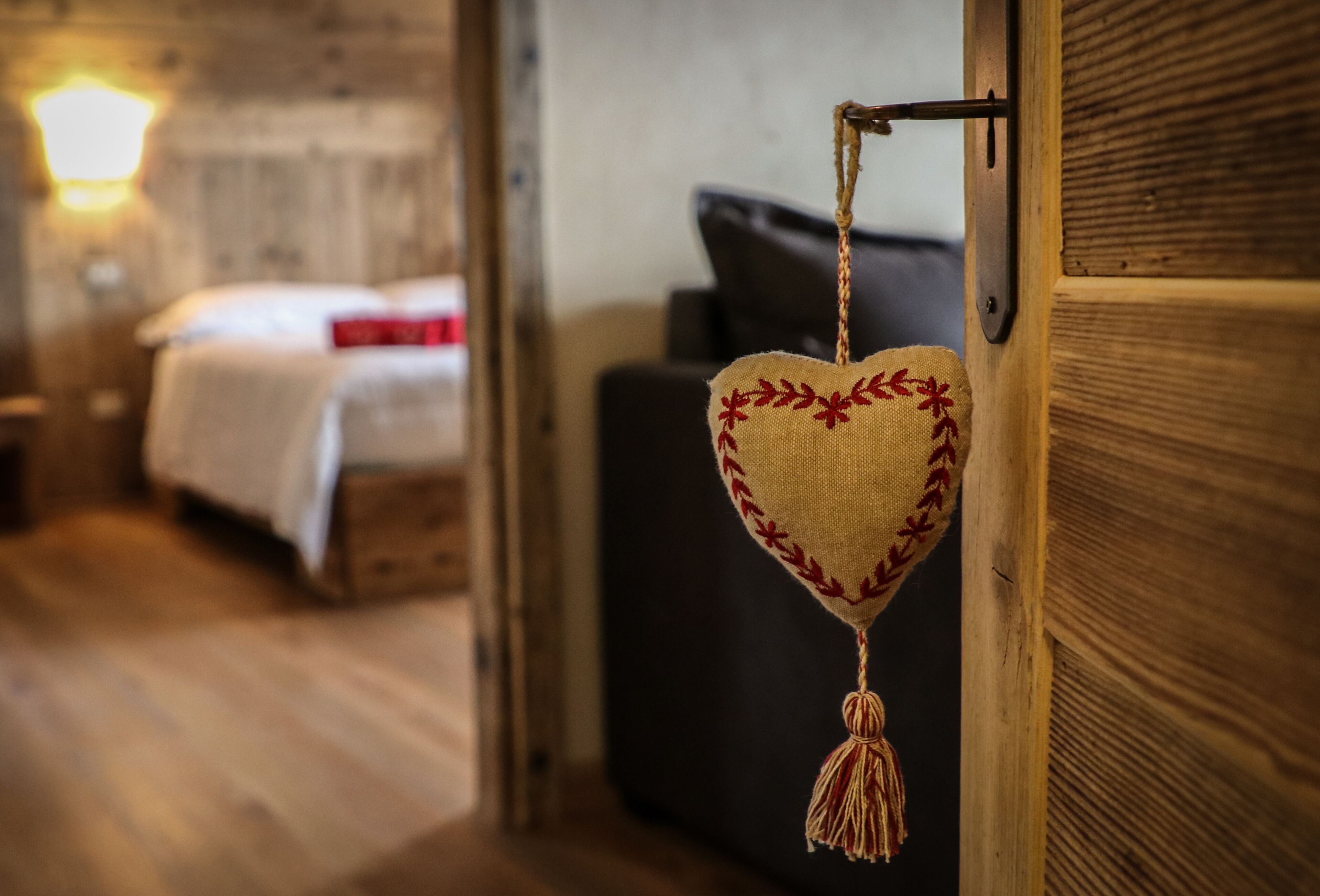 A heart-shaped hanging decoration with tassels, a wooden door, and a bed with a white blanket and red pillow in the background.