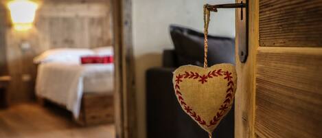 Decoración colgante en forma de corazón con flecos, una puerta de madera y una cama con manta blanca y almohada roja al fondo.
