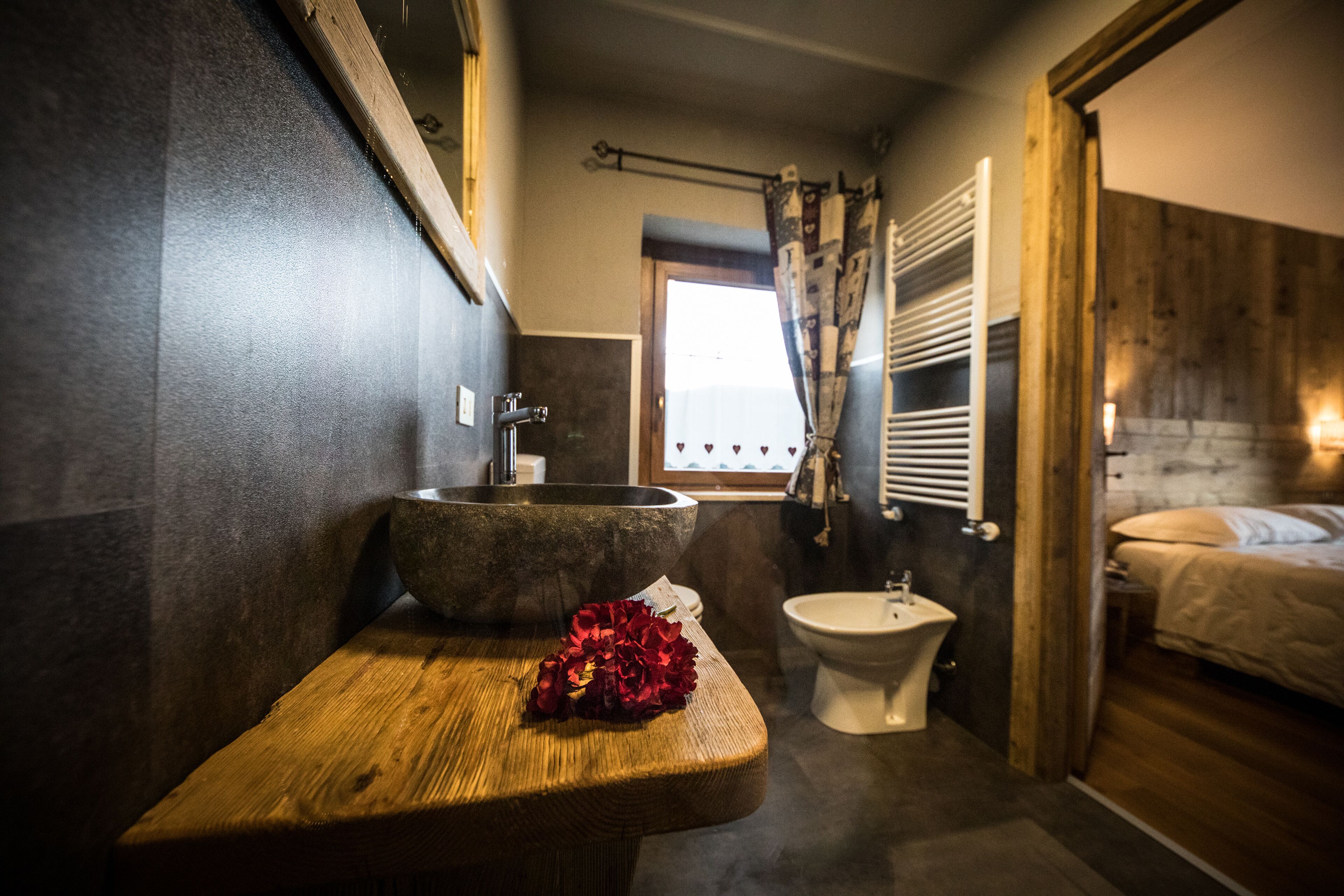 A bathroom with a wooden bench, a stone basin, a white toilet, and a bed with a wooden headboard.