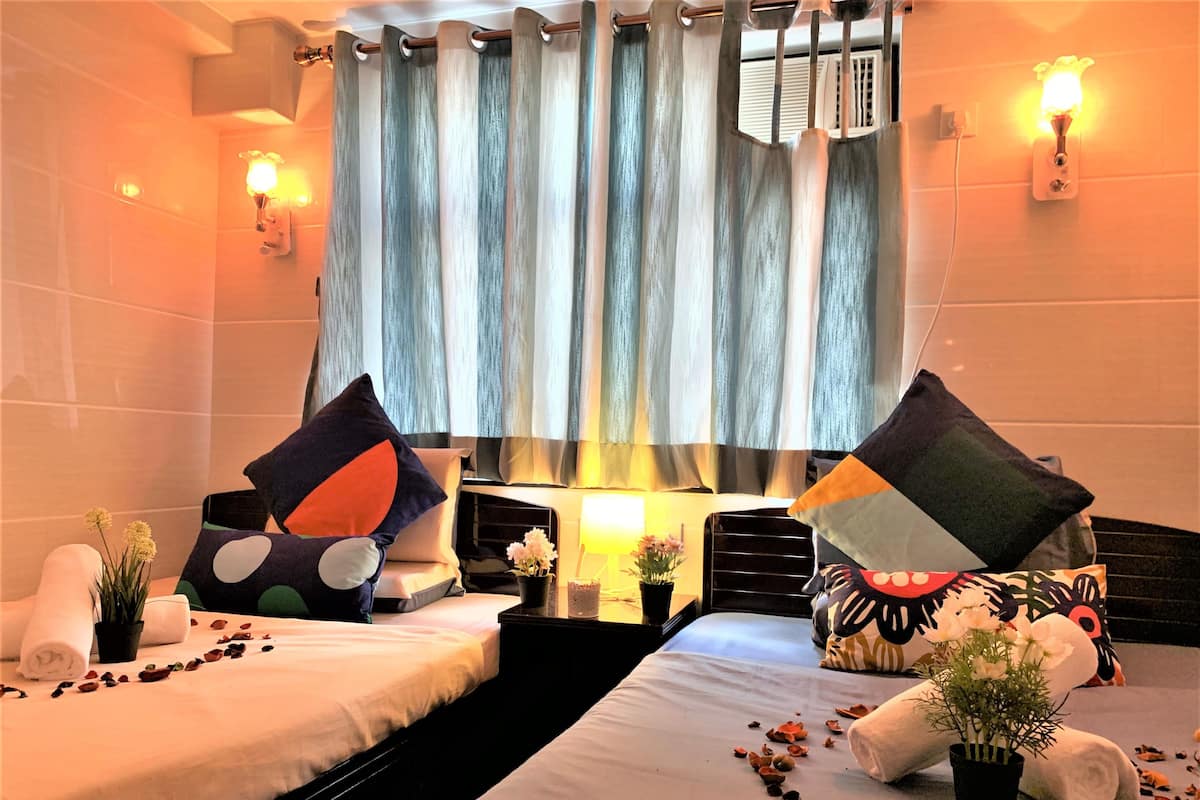A hotel room with two beds, each with a pillow and a small plant. There is a window with curtains, a bedside table with a lamp, and a wall-mounted air conditioner.